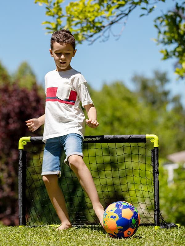 Junge in Freizeitkleidung spielt mit buntem Fußball vor kleinem Tor im Garten.