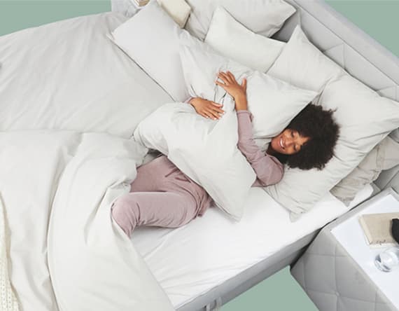 Frau in rosa Schlafanzug kuschelt sich lächelnd in ein Bett mit weißen Kissen und Bettdecken