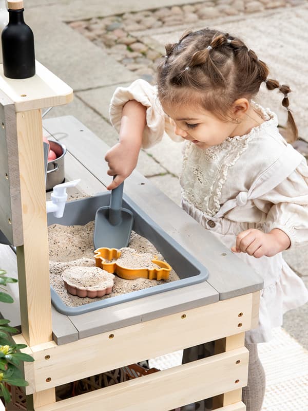Kind spielt draußen mit einer Schaufel im Sandkasten, auf einem Holztisch stehen Förmchen und ein Spielzeug.