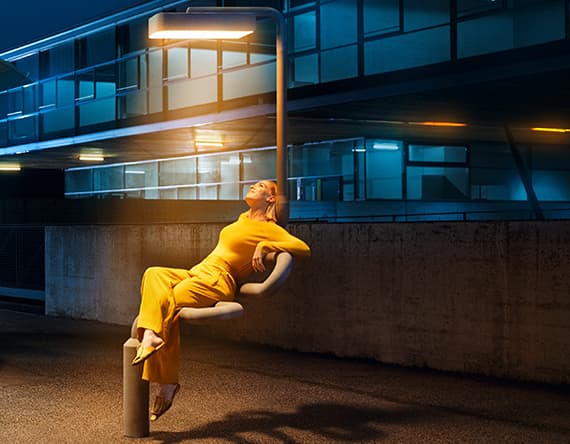 Frau in gelbem Outfit lehnt sich nachts an eine Straßenlaterne und blickt nach oben. Moderne Architektur im Hintergrund.