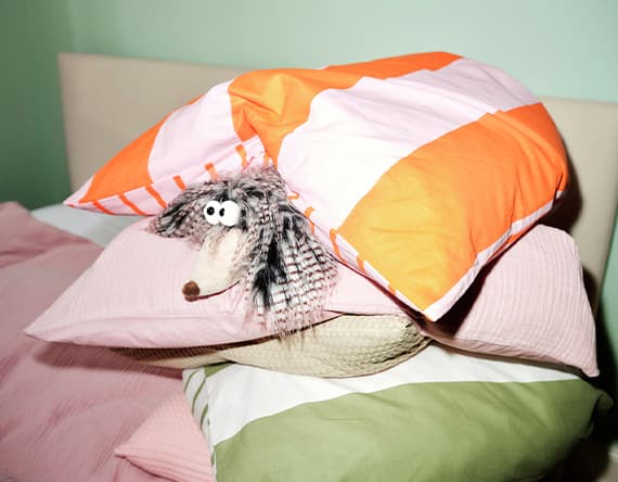 Plüschigel mit großen Augen unter gestapelten, bunten Kissen im Bett versteckt