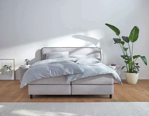 Modernes Bett mit grauer Bettwäsche, umgeben von Nachttischen, einer Zimmerpflanze und einem gerahmten Bild.