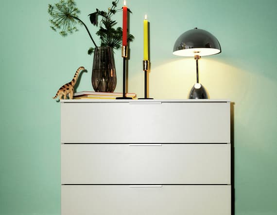 Weiße Kommode mit Lampe, Kerzen, Vase und Spielzeugdinosaurier davor grüner Wand.