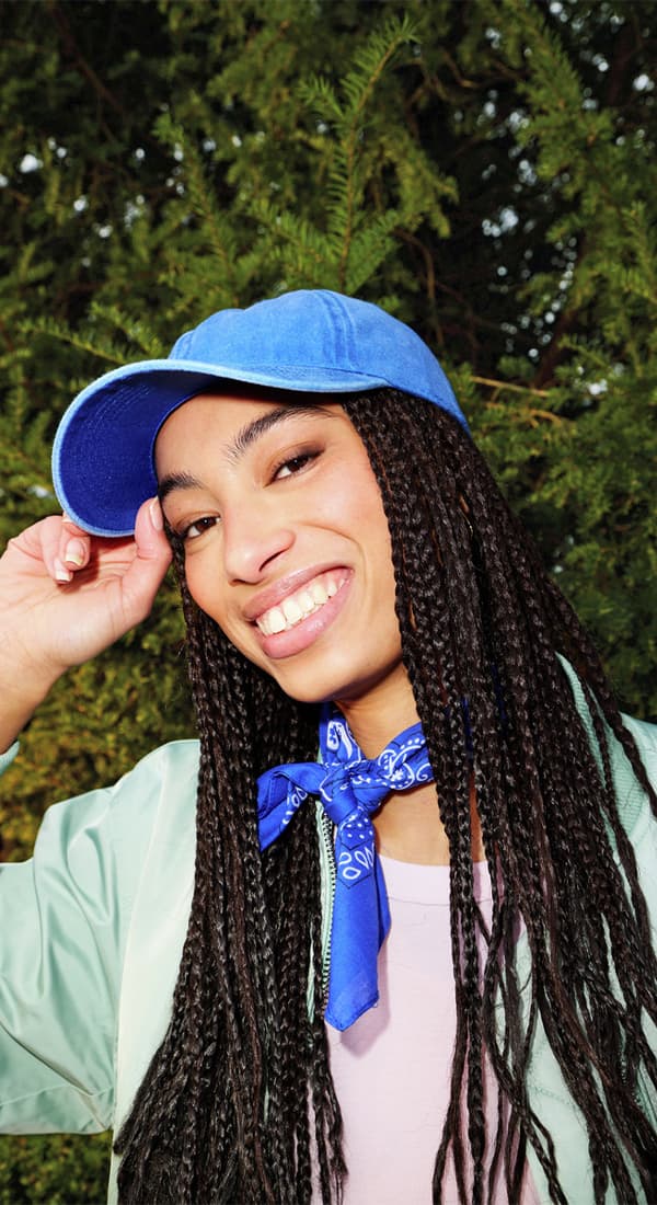 Person mit langen Zöpfen im Park, trägt blaue Cap, mintgrüne Jacke und blaues Halstuch, Hand am Schild der Cap