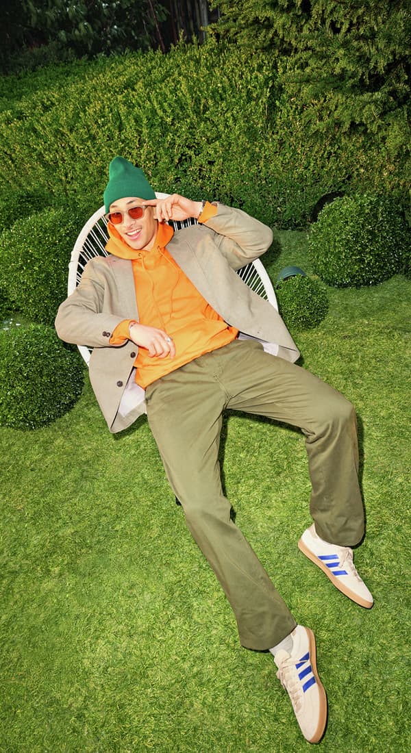 Mann in orange Hoodie, Sakko und grüner Mütze liegt entspannt auf Stuhl im Garten, Füße in Sneakers nach vorn ausgestreckt