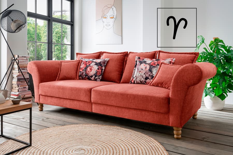 Rotes Sofa mit geblümten Kissen in hellem Wohnzimmer, links Bücherstapel, rechts große Zimmerpflanze