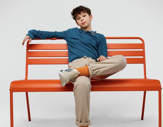 Junge sitzt lässig auf orangener Bank, ein Bein überschlagen, trägt blaues Langarmshirt, helle Hose und Sneaker