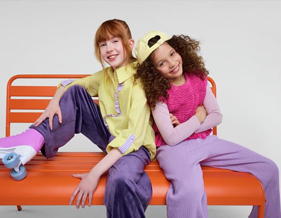 Zwei Kinder in bunten Outfits sitzen lässig auf einer orangefarbenen Bank, eines trägt Rollschuhe