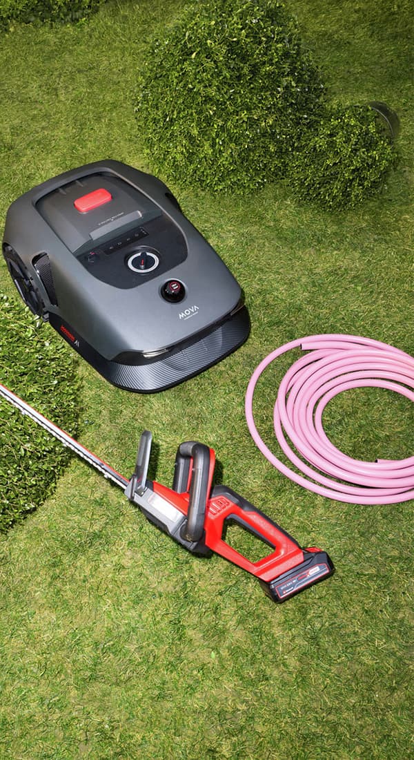 Rasenroboter, Heckenschere und aufgerollter Gartenschlauch liegen auf kurz gemähtem Rasen neben Formschnitt-Sträuchern