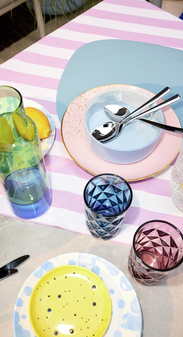 Gedeckter Tisch mit gestreifter Tischdecke, bunten Tellern, Gläsern und Schalen, zwei Löffel liegen in einer Schale