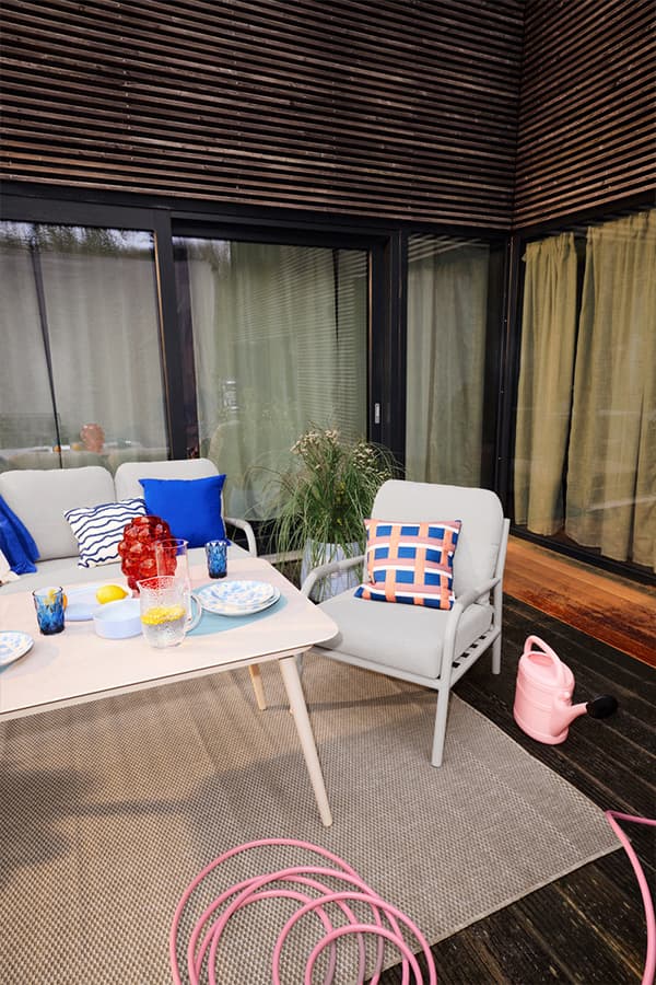 Gedeckter Terrassentisch mit bunten Gläsern, Sofa und Sessel mit Kissen, daneben Gießkanne und aufgerollter Gartenschlauch