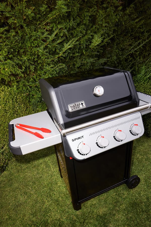 Gasgrill Weber Spirit im Garten, mit geschlossenem Deckel, seitlicher Ablage und Grillzange auf Rasen vor Hecke