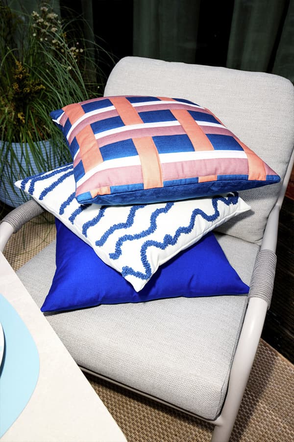 Drei bunte Zierkissen mit Muster liegen gestapelt auf einem hellen Sessel auf der Terrasse