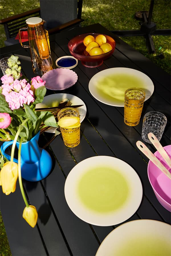 Sommerlich gedeckter Gartentisch mit buntem Geschirr, Karaffe, Gläsern mit Saft, Schale mit Zitronen und Blumenstrauß