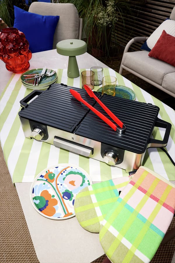 Elektrogrill mit roten Grillzangen auf gedecktem Gartentisch mit bunten Tellern, Gläsern und karierten Ofenhandschuhen