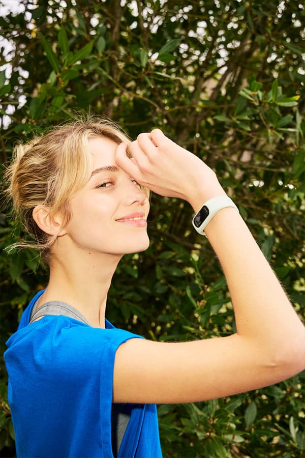 Person im blauen Sportoberteil mit Fitnessarmband hebt den Arm vor grünem Busch im Freien