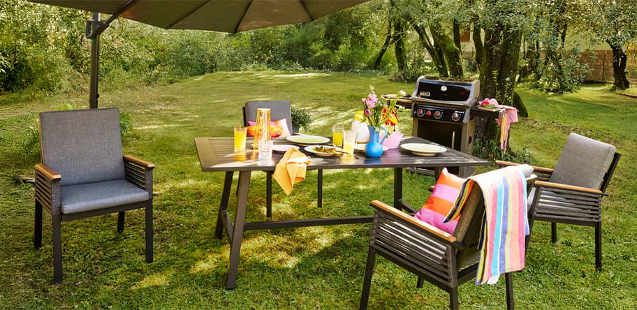 Gartentisch mit vier Stühlen, gedeckter Frühstückstafel und Grill unter Sonnenschirm auf grüner Wiese