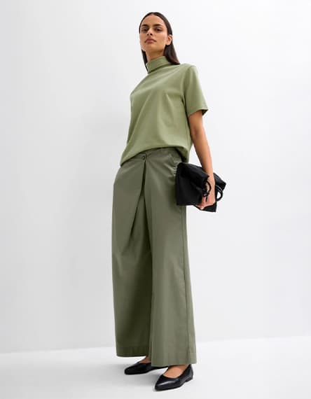 Frau in olivgrünem Oberteil und weit geschnittener olivgrüner Hose, hält schwarze Tasche, trägt schwarze Schuhe.