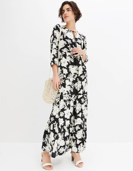 Frau in langem geblümtem Kleid mit 3/4-Ärmeln, Basttasche über der Schulter und offenen Sandalen