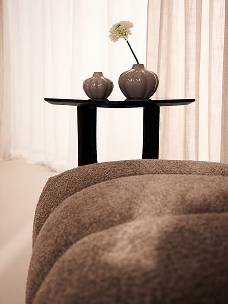 Kleiner Beistelltisch mit zwei runden Vasen und einer Blume hinter einer strukturierten Sitzfläche vor hellen Vorhängen