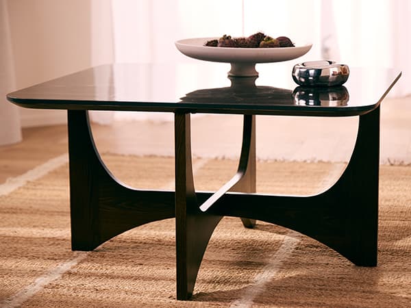 Quadratischer Couchtisch mit geschwungenem Fußgestell, darauf flache Schale mit Deko und runde Dose auf Teppich