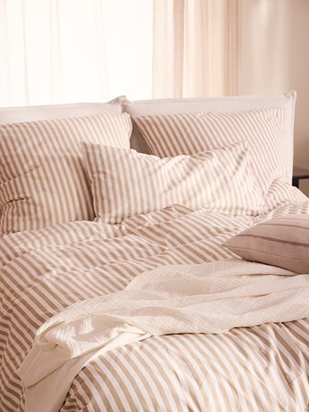 Beige-weiß gestreifte Bettwäsche auf Doppelbett mit mehreren Kissen und locker darüberliegendem Plaid