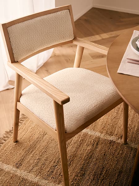 Heller Esszimmerstuhl mit gepolsterter Sitzfläche und Rückenlehne neben rundem Holztisch auf gewebtem Teppich