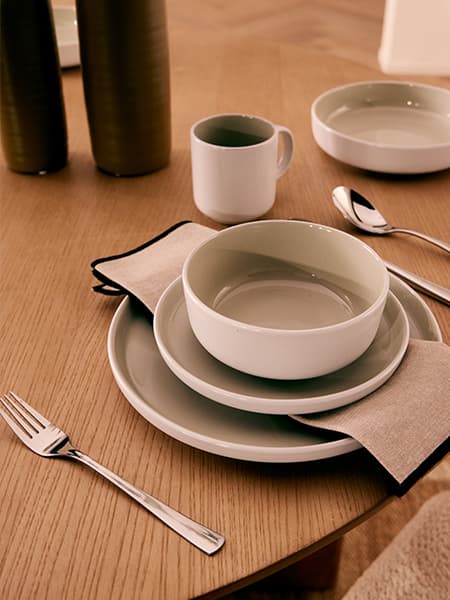 Tischgedeck mit gestapelten Tellern, Schale und Stoffserviette, daneben Besteck, Tasse und Schale auf Holzoberfläche
