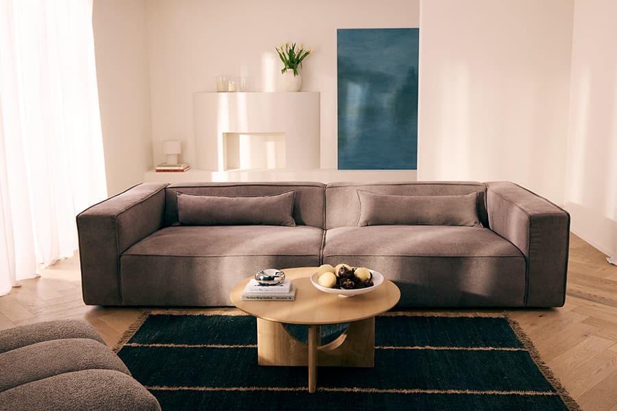 Großes graues Sofa vor heller Wand, runder Couchtisch mit Obstschale und Büchern, dahinter Kamin und blaues Wandbild