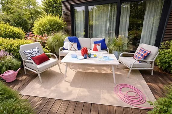 Sitzgruppe auf Terrasse mit Sofa, zwei Sesseln, Tisch mit Geschirr, bunten Kissen und Gartenschlauch vor bodentiefen Fenstern
