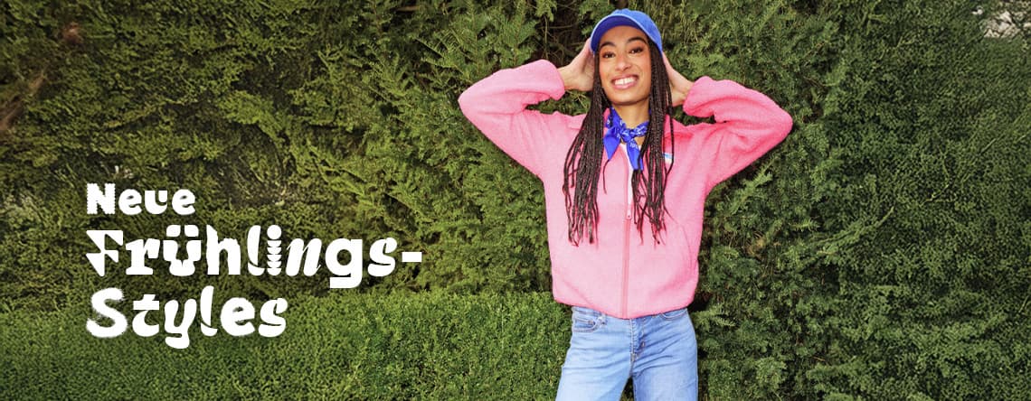 Neue Frühlings-Styles – Person in pinker Jacke und Jeans steht mit erhobenen Armen vor grüner Hecke