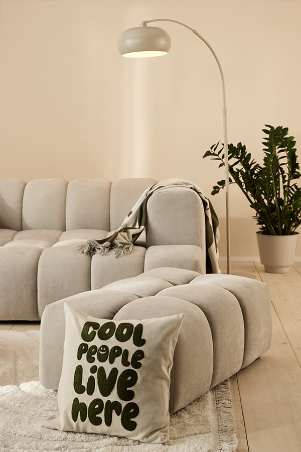 Kissen mit Schrift „Cool people live here“ vor beigem Sofa, Hocker, Stehlampe und Zimmerpflanze im Wohnzimmer