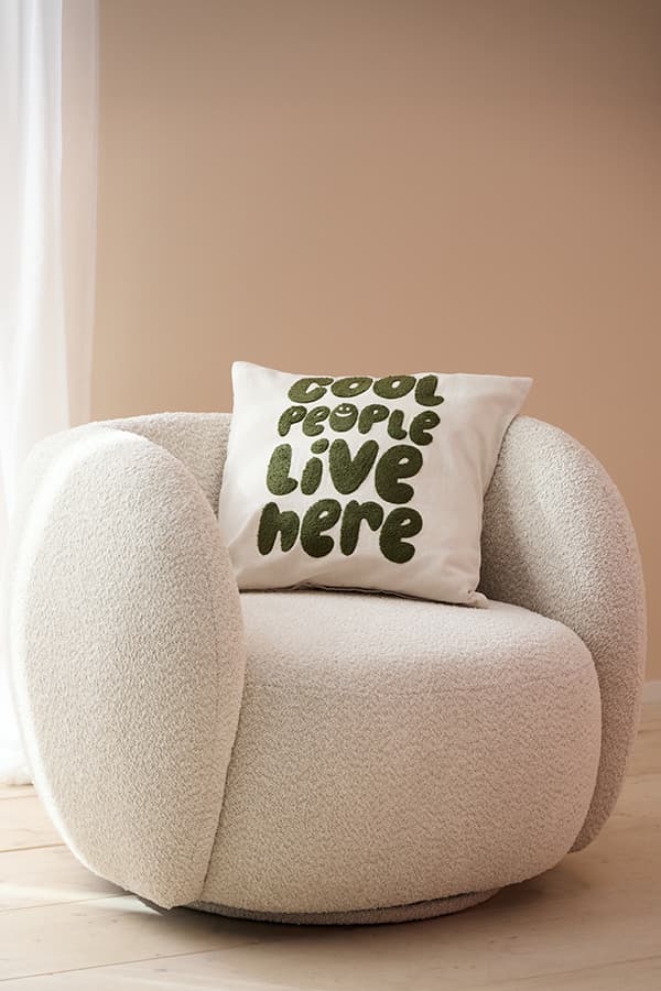 Kissen mit Aufdruck „Cool people live here“ auf rundem, cremefarbenem Sessel vor beigefarbener Wand