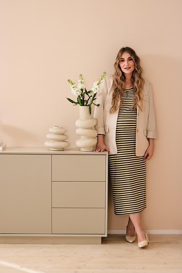 Frau in gestreiftem Kleid und hellem Blazer lehnt an Sideboard mit Deko-Vasen und Blumen in modernem Wohnzimmer