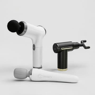 Drei Massagegeräte in Weiß und Schwarz mit unterschiedlichen Aufsätzen stehen und liegen nebeneinander auf hellem Untergrund