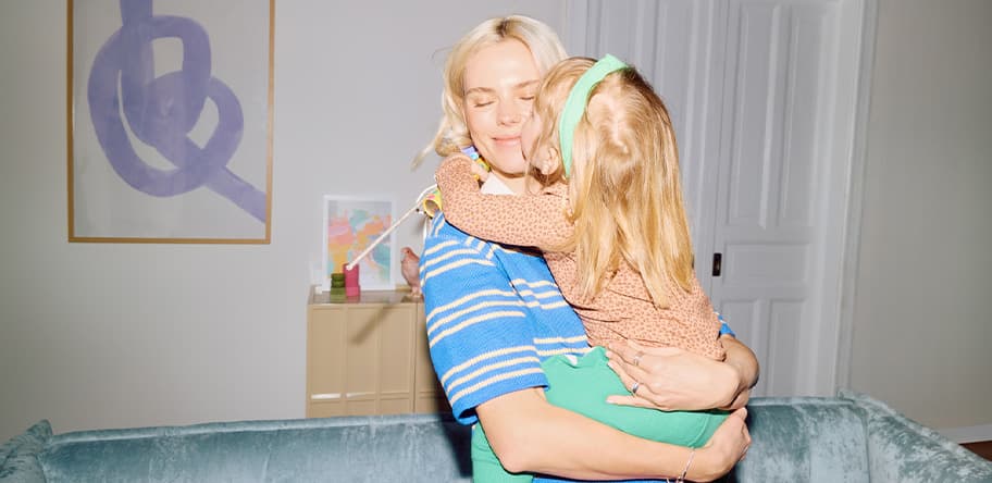 Erwachsene Person im gestreiften Oberteil hält kleines Mädchen mit Haarband im Arm in einem Wohnzimmer