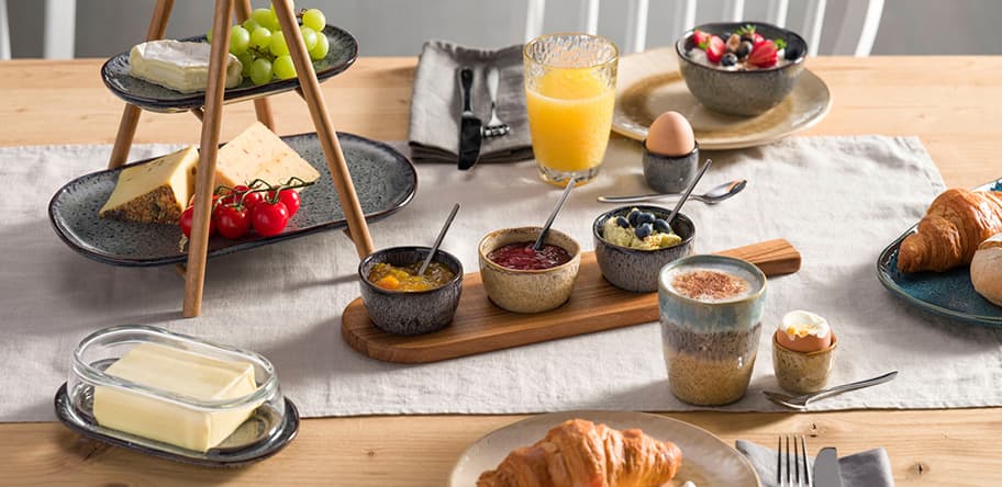 Frühstückstisch mit Croissants, Käse, Obst, Marmeladen, Eiern, Butter und Saft auf Holz- und Keramikgeschirr