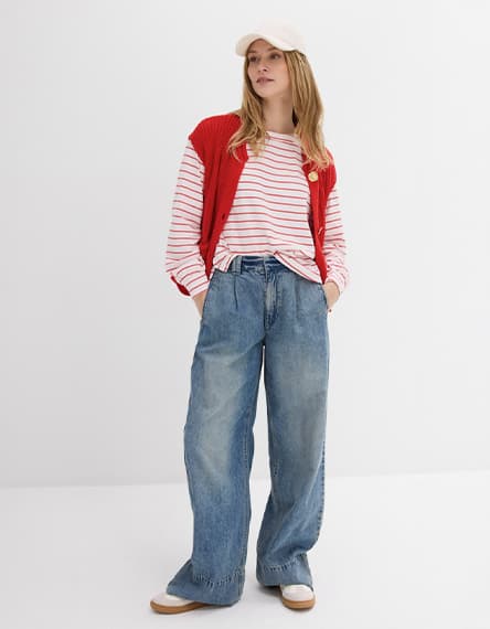 Person mit rotem Cardigan über gestreiftem Langarmshirt und weitem Jeanslook, trägt Basecap und steht mit Händen in den Taschen