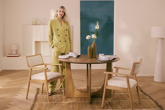 Frau im hellen Anzug steht an rundem Holztisch mit zwei Stühlen, Geschirr und Vasen mit Blumen im modernen Esszimmer