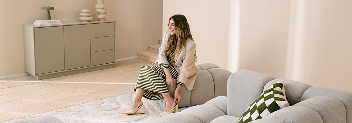 Frau sitzt auf hellem Sofa, richtet ihre Schuhe, im Hintergrund Sideboard und Teppich im modernen Wohnzimmer