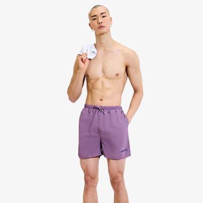 Mann in lila Badeshorts steht frontal, hält ein kleines Handtuch an der Schulter und hat eine Hand in der Hosentasche