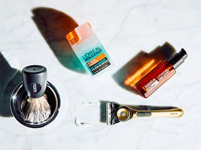 Rasierpinsel, Rasierer und zwei L’Oréal Men Expert Pflegeprodukte auf hellem Marmoruntergrund