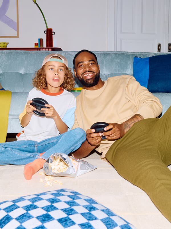 Erwachsener und Kind sitzen vor dem Sofa, spielen mit Controllern Videospiel, offene Popcorntüte auf dem Teppich