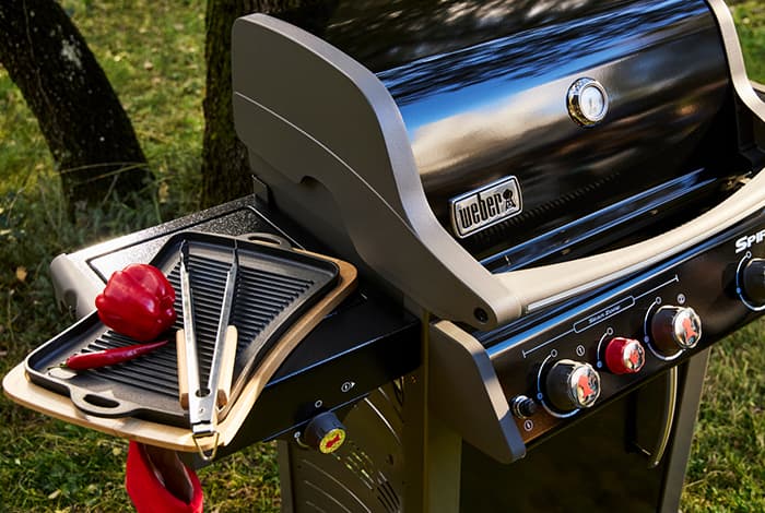 Gasgrill im Garten mit seitlicher Ablage, Grillplatte, Grillzange und roter Paprika neben den Bedienknöpfen