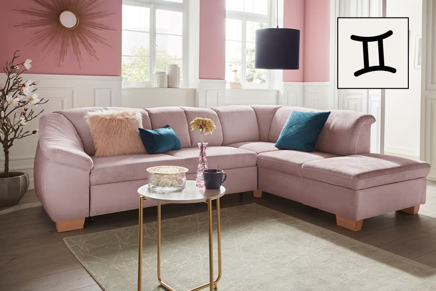 Ecksofa in L-Form in rosa Wohnzimmer, mit Zierkissen, kleinem runden Tisch und Zwillinge-Sternzeichen-Symbol an der Wand