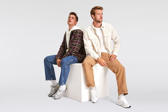 Zwei Männer sitzen auf weißem Block, einer in Karohemd und Jeans, der andere in heller Jacke und beigefarbener Hose