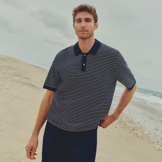 Mann am Strand in dunkelblau gestreiftem Poloshirt, eine Hand in der Hosentasche, Meer im Hintergrund