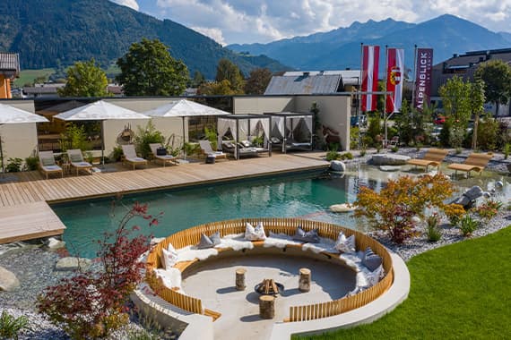 Runde Feuerstelle mit Sitzkissen am Naturpool, dahinter Liegen und Pavillons, Berge im Hintergrund