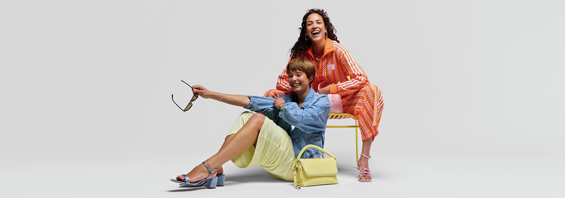 Zwei Frauen in farbenfrohen Outfits, eine sitzt mit Sonnenbrille und Handtasche am Boden, die andere steht dahinter auf einem Stuhl