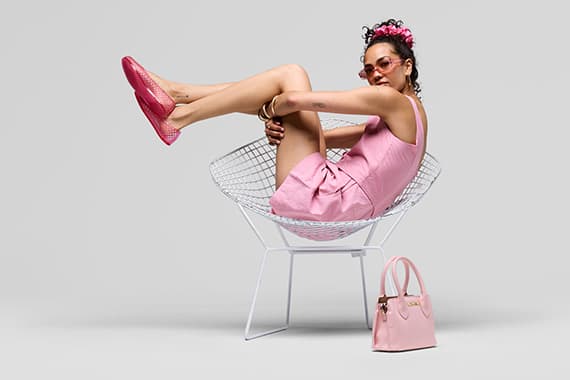 Frau in rosa Kleid sitzt eingerollt im Designerstuhl, trägt passende Schuhe und daneben steht eine rosa Handtasche